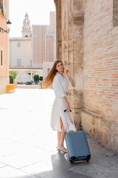 Piena lunghezza di donna rossa felice in piedi con i bagagli sulla strada vicino alla cattedrale di Valencia — Foto stock