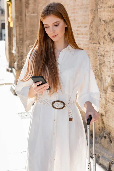 Jeune rousse femme tenant poignée de bagages et en utilisant smartphone dans la rue en Europe — Photo de stock