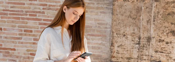 Jovem ruiva mulher usando smartphone na rua europeia perto da parede de tijolo, banner — Fotografia de Stock