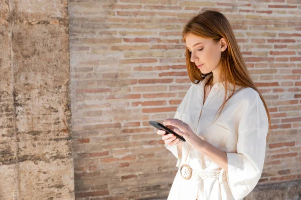 Young redhead woman messaging on smartphone near brick wall on urban street — Stock Photo