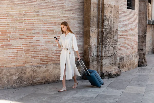 Full length of redhead woman walking with baggage and using smartphone on european street — Stock Photo