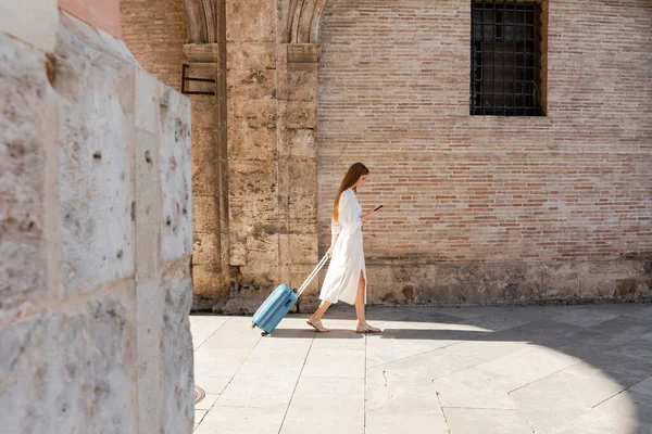 Vista lateral de mujer pelirroja caminando con equipaje y usando teléfono inteligente en la calle europea - foto de stock