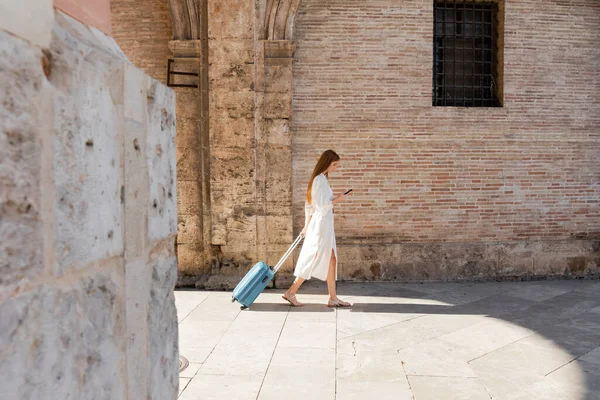 Side view of redhead woman walking with baggage and using smartphone on european street — Stock Photo