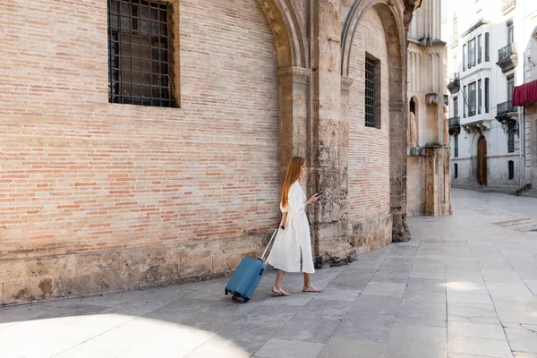 Comprimento total da mulher ruiva andando com bagagem e usando smartphone em valência — Fotografia de Stock