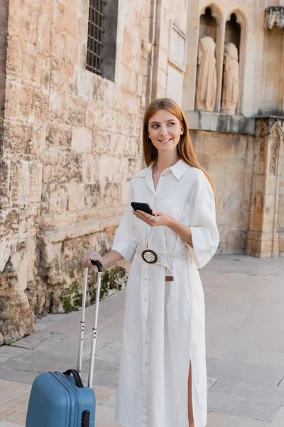 Heureuse rousse femme tenant poignée de bagages et en utilisant smartphone sur la rue de valencia — Photo de stock