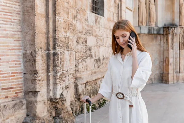 Donna rossa sorridente che tiene in mano la maniglia dei bagagli e parla sullo smartphone in valencia — Foto stock