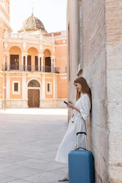 Pleine longueur de femme rousse gaie debout près de la valise et en utilisant un smartphone sur la rue de Valencia — Photo de stock