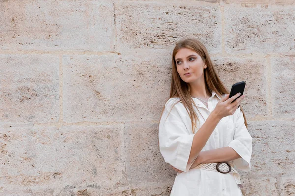 Redhead woman holding smartphone while standing near wall on street of valencia — Stock Photo