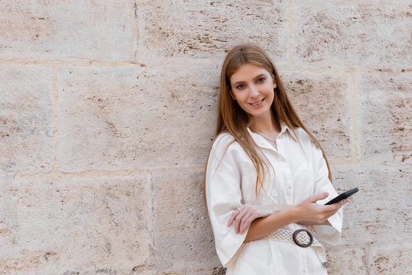 Cheerful redhead woman holding smartphone while standing near wall on street of valencia — Stock Photo