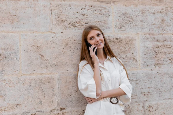 Cheerful redhead woman talking on smartphone while standing near wall on street of valencia — Stock Photo