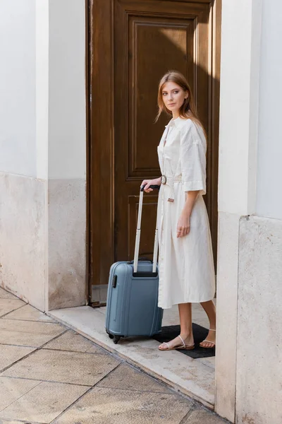 Full length of redhead woman in white dress holding luggage handle and standing near door on street in valencia — Stock Photo