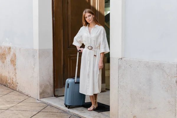 Повна довжина рудої жінки в білій сукні, що стоїть з валізою біля дверей на європейській вулиці — стокове фото