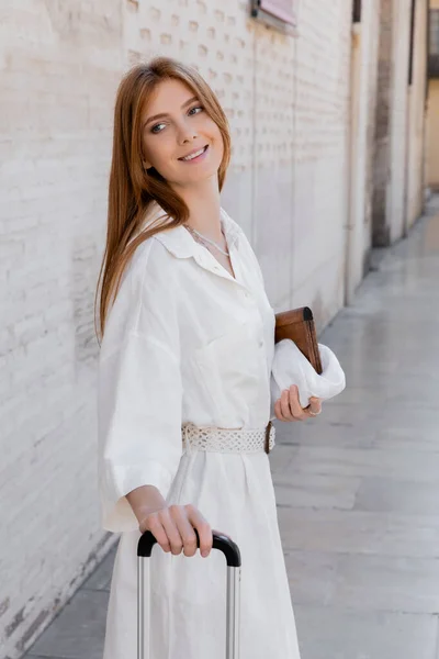 Cheerful woman in dress holding luggage handle and standing on white street in valencia — Stock Photo