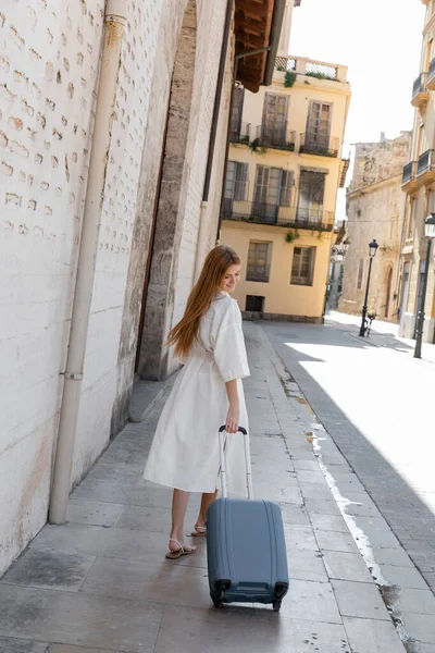 Jeune femme souriante en robe de marche avec valise dans la rue à Valencia — Photo de stock