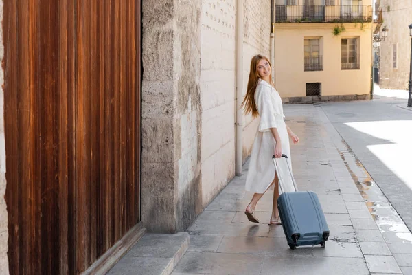 Young cheerful woman in dress walking with suitcase on street in valencia — Stock Photo