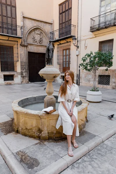 Comprimento total da mulher ruiva em vestido branco sentado perto da fonte na rua de valencia — Fotografia de Stock