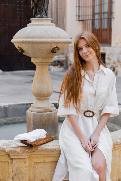 Smiling redhead woman in white dress sitting near fountain on street of valencia — Stock Photo