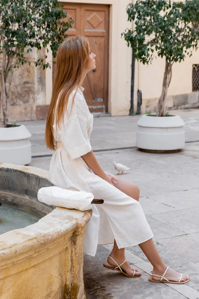 Vista laterale di donna rossa in abito bianco seduta vicino fontana sulla strada di valencia — Foto stock