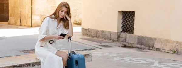 Rothaarige Frau im Kleid sitzt neben Koffer und benutzt Handy in valencia, Banner — Stockfoto