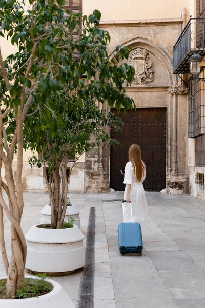 Вид сзади рыжеволосой женщины в платье, стоящей с рюкзаком и держащей смартфон на древней улице Валенсии — Stock Photo