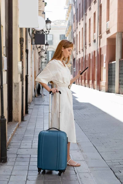 Redhead woman in dress using smartphone and standing with luggage on urban street in valencia — Stock Photo