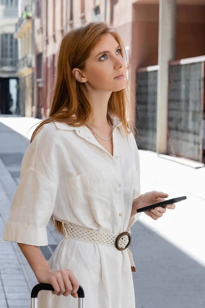 Redhead woman in dress holding luggage handle and smartphone on urban street in valencia — Stock Photo