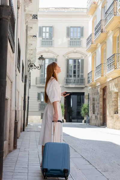 Рыжая женщина в платье, держащая смартфон и стоящая с чемоданом на городской улице в Валенсии — стоковое фото