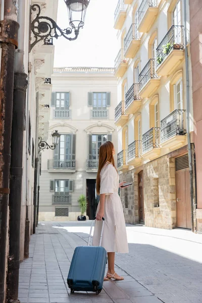 Redhead tourist in dress holding smartphone and standing with suitcase on urban street in valencia — Stock Photo