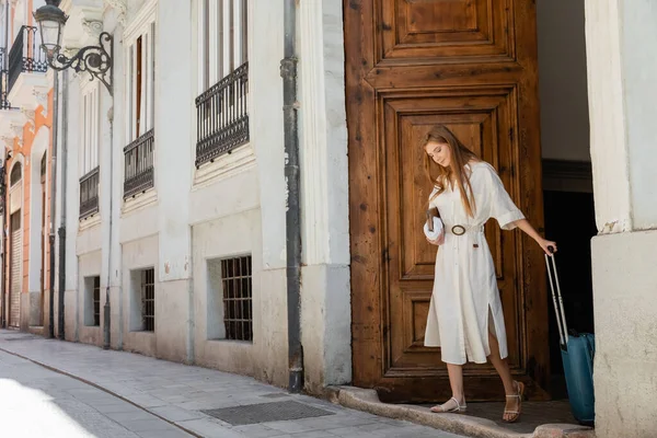 Mulher ruiva segurando bolsa e mala perto da porta de entrada de madeira em valencia — Fotografia de Stock