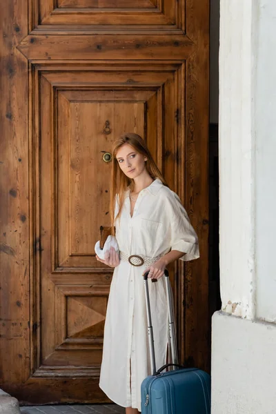 Young woman holding purse and luggage near wooden entrance door in valencia — Stock Photo