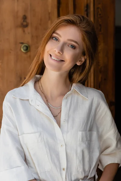 Portrait of smiling redhead woman looking at camera near wooden door — Stock Photo