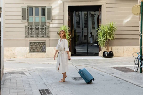 Full length of happy woman in dress walking with luggage on european street in Valencia — Stock Photo