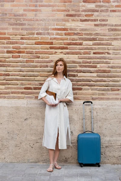 Longitud completa de mujer pelirroja en vestido sosteniendo bolso y de pie con la maleta cerca de la pared de ladrillo en la calle europea - foto de stock