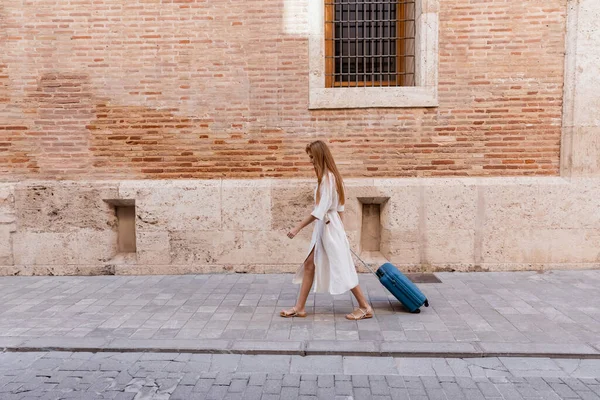 Piena lunghezza della donna rossa in abito camminare con valigia vicino muro di mattoni sulla strada europea — Foto stock