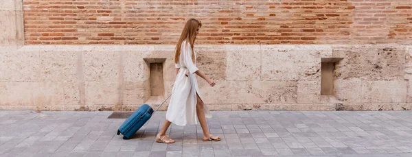 Piena lunghezza della donna rossa in abito camminare con valigia vicino muro di mattoni sulla strada europea, banner — Foto stock