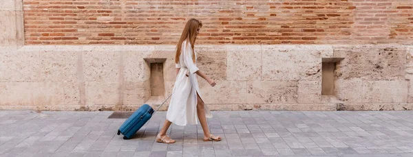 Full length of redhead woman in dress walking with suitcase near brick wall on european street, banner — Stock Photo