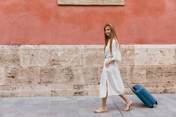 Piena lunghezza di donna rossa in abito camminare con valigia vicino muro di mattoni sulla strada europea in Valencia — Foto stock