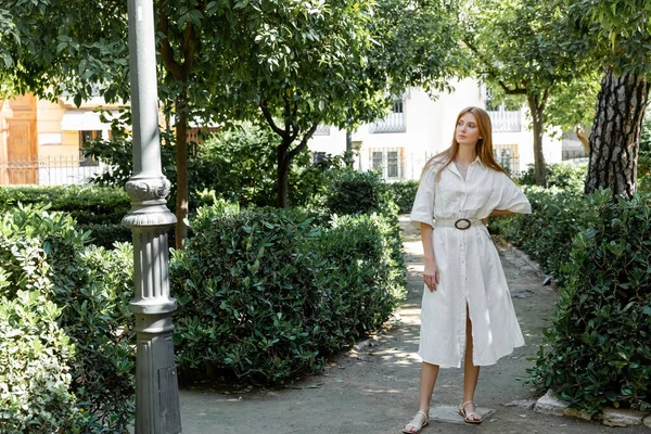 Pleine longueur de jeune rousse femme en robe debout avec la main sur la hanche sur la rue verte à Valencia — Photo de stock