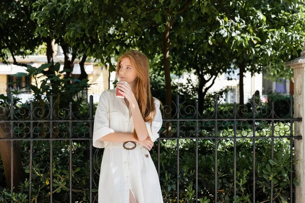 Young redhead woman in dress holding coffee to go near fence in european green park — Stock Photo