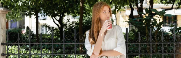 Young redhead woman in dress holding coffee to go near fence in european green park, banner — Stock Photo