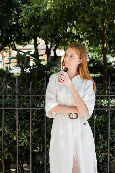 Jeune rousse femme en robe blanche tenant café pour aller près de la clôture dans le parc vert européen — Photo de stock