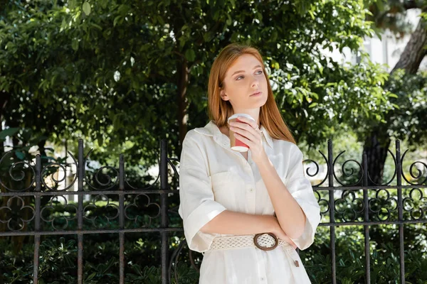 Thoughtful redhead woman in white dress holding coffee to go near fence in european green park — Stock Photo