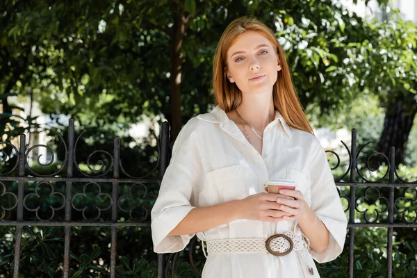 Rousse femme en robe blanche tenant café pour aller et regardant la caméra près de la clôture dans le parc vert européen — Photo de stock