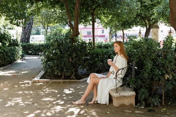 Longitud completa de mujer pelirroja en vestido sosteniendo café para ir y sentado en banco de hormigón en el parque verde europeo - foto de stock