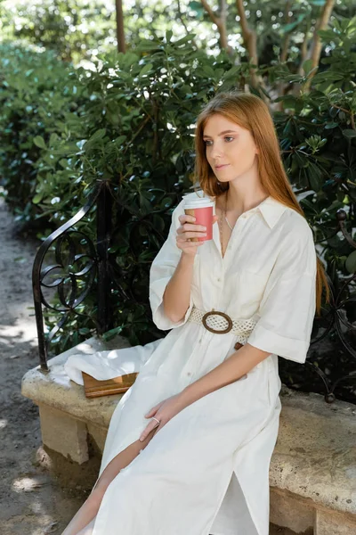 Young redhead woman in dress holding paper cup and sitting on concrete bench in green park — Stock Photo