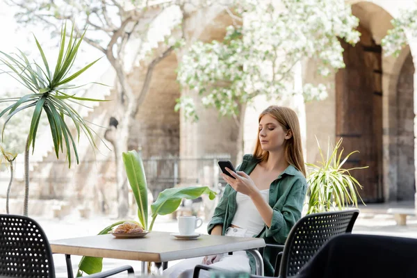 Rothaarige Frau hält Smartphone neben Tasse und Croissant auf Tisch in Café-Terrasse — Stockfoto