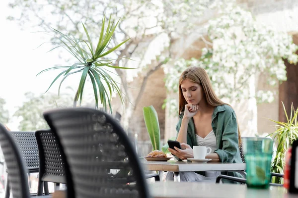 Рыжая женщина с помощью смартфона возле чашки кофе и круассана на столе на террасе кафе — стоковое фото