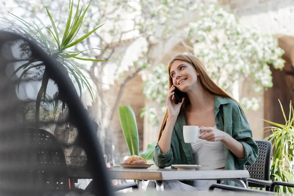 Mujer feliz hablando en el teléfono inteligente y la celebración de la taza de café cerca de croissant en la mesa en la terraza del café - foto de stock