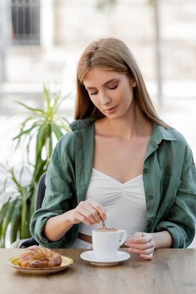 Redhead woman stirring coffee with spoon near croissant on table in cafe terrace — Stock Photo