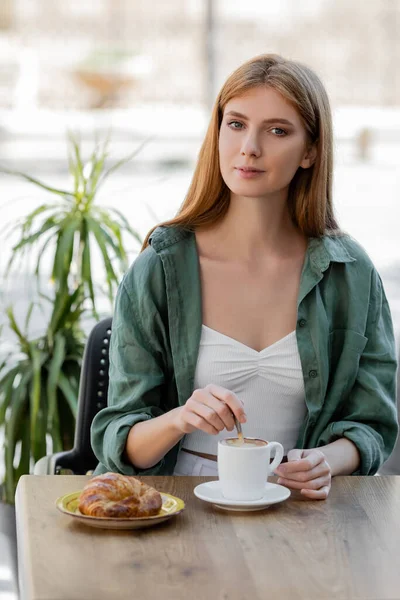 Redhead woman stirring coffee with spoon and looking at camera on summer terrace — Stock Photo